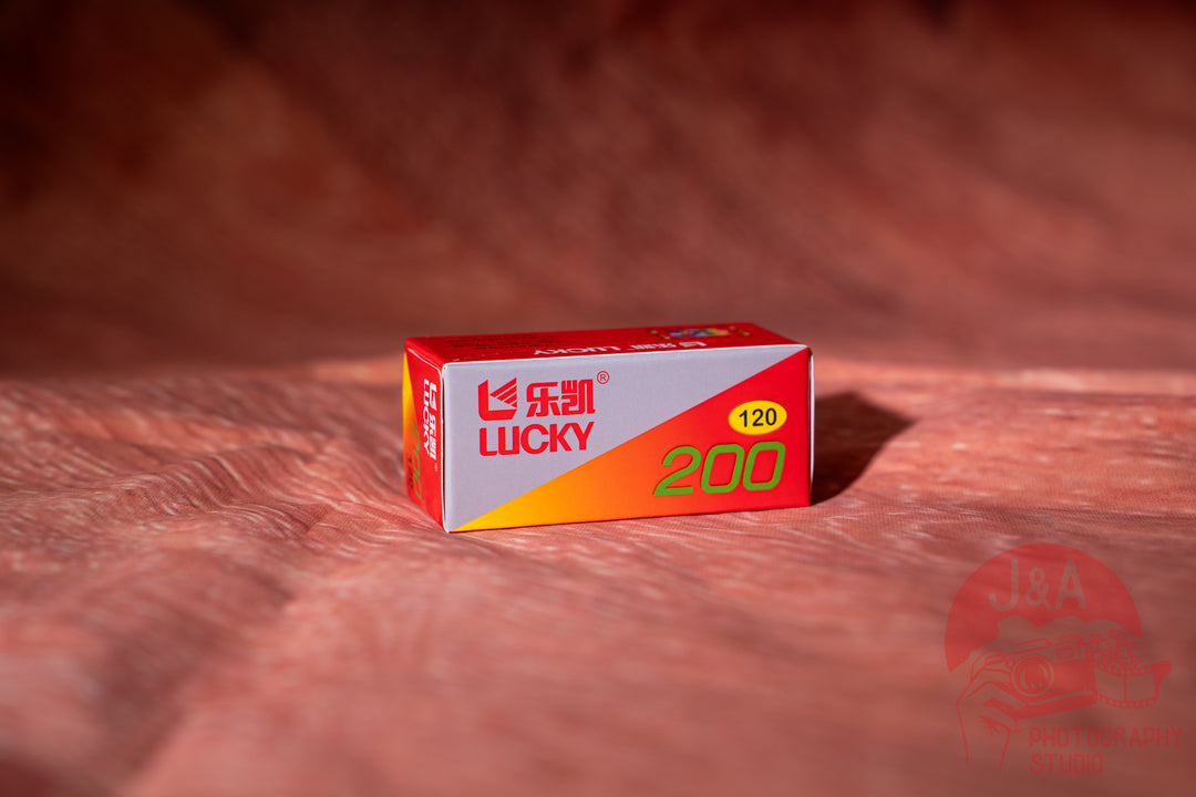 A small red, white, and orange box of Lucky 200 film stands on a textured reddish-brown surface with a matching blurred background. The box features the red and white Lucky brand logo and a yellow circle indicating 120.