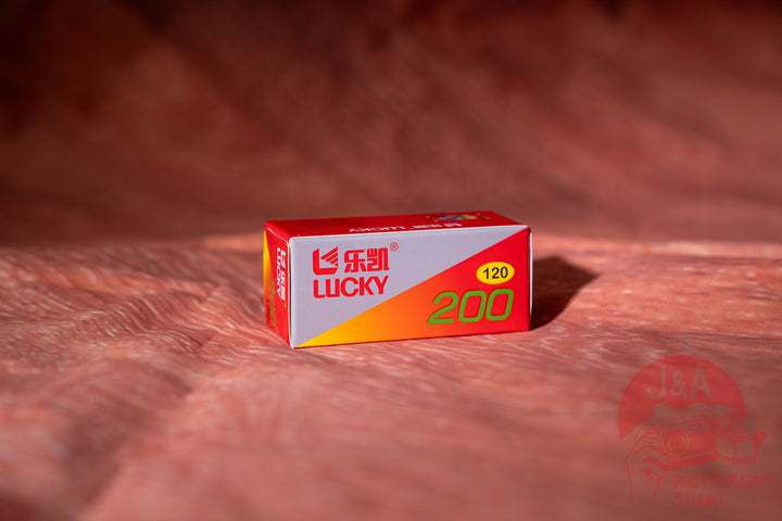 A small red, white, and orange box of Lucky 200 film stands on a textured reddish-brown surface with a matching blurred background. The box features the red and white Lucky brand logo and a yellow circle indicating 120.