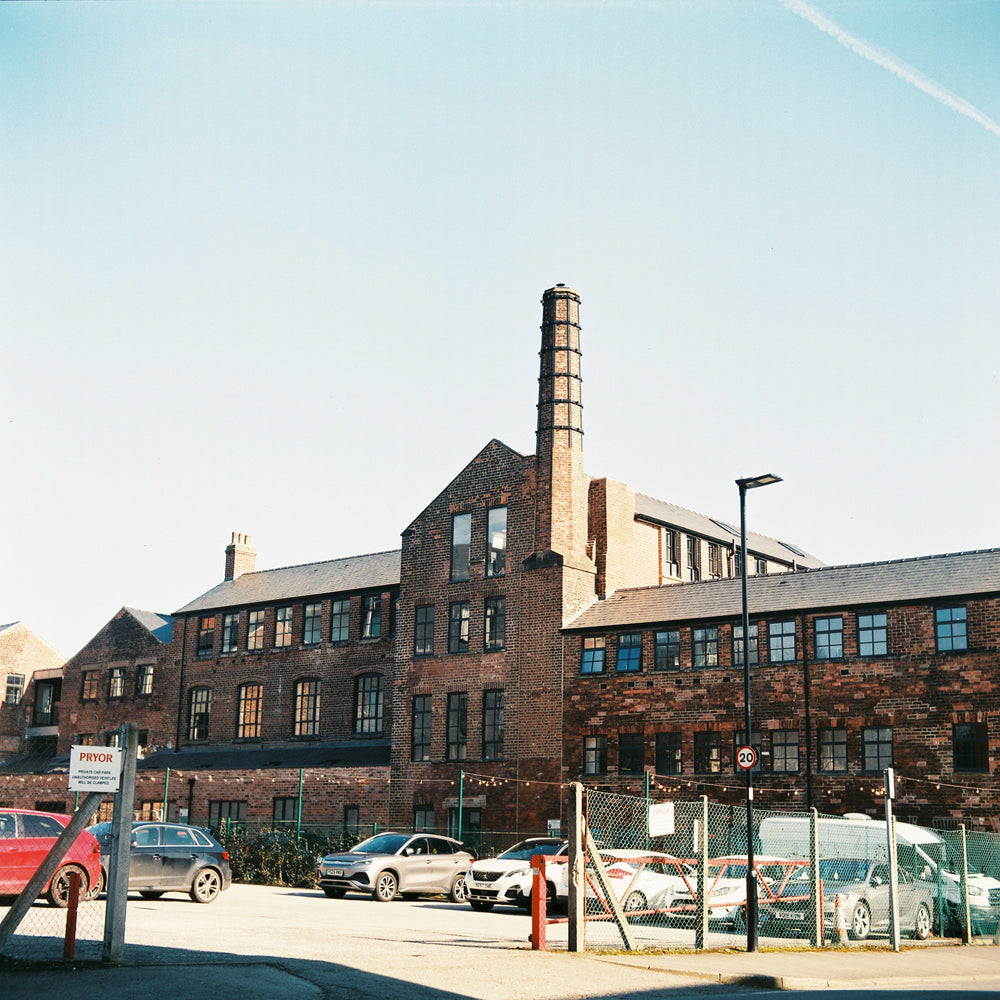 A large brick industrial factory building with a tall chimney and multiple windows, photographed outdoors under a pale blue sky. Several cars are parked in front near a chain-link fence, with a street sign and lamp visible.