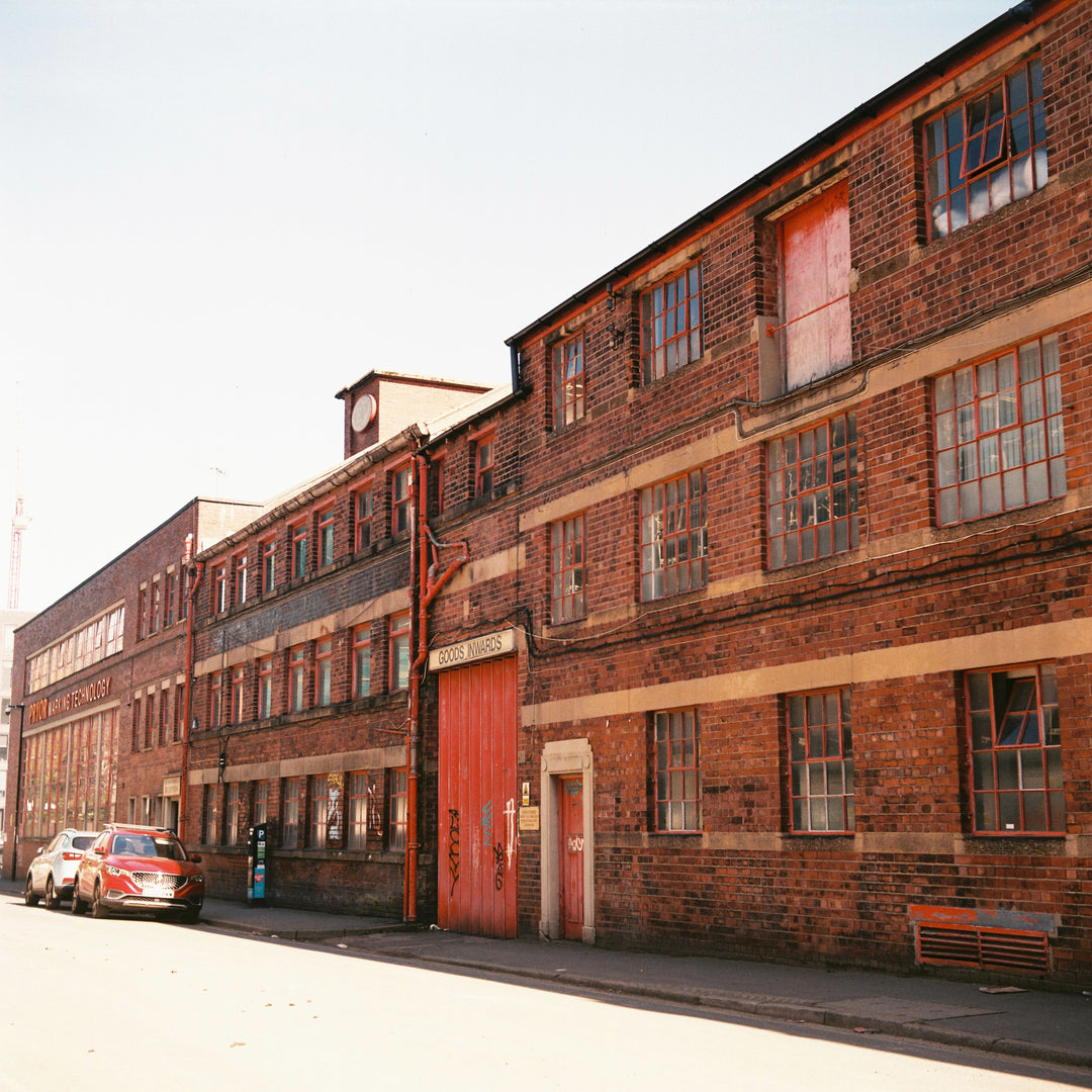 A long row of old brick industrial buildings with large windows, a red door, parked cars along the street, and a pale sky above.