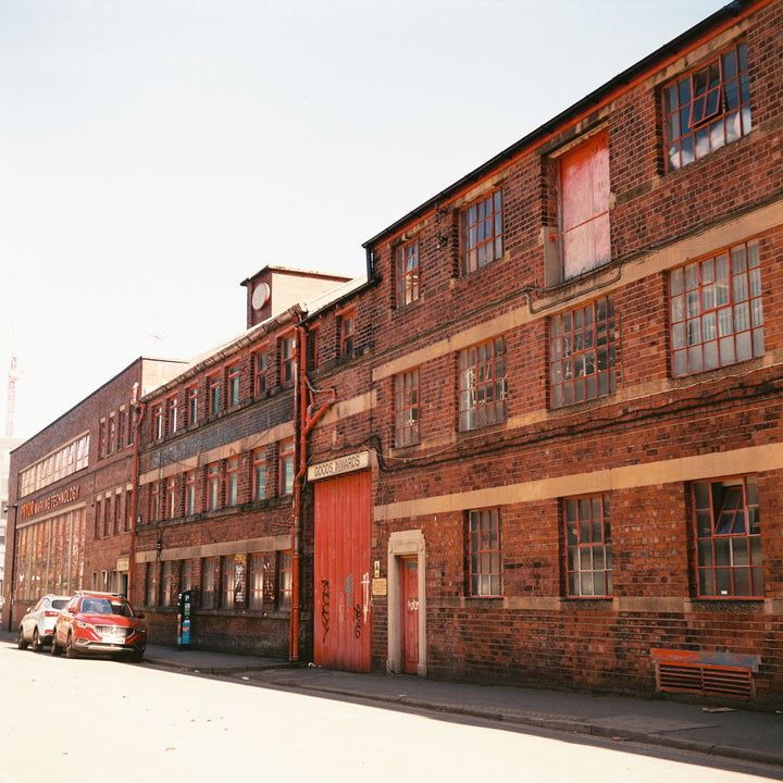 A long row of old brick industrial buildings with large windows, a red door, parked cars along the street, and a pale sky above.